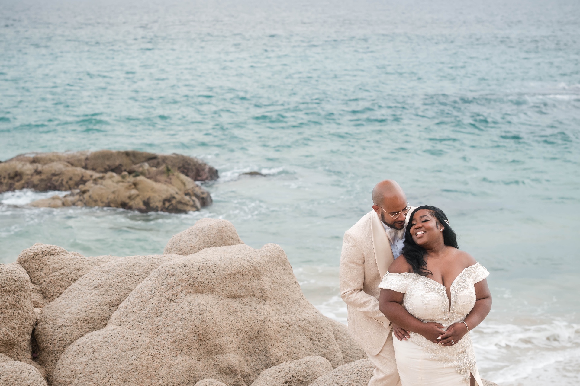 Couple embrace on beach rocks Los Cabos Mexico destination wedding cream suit ocean