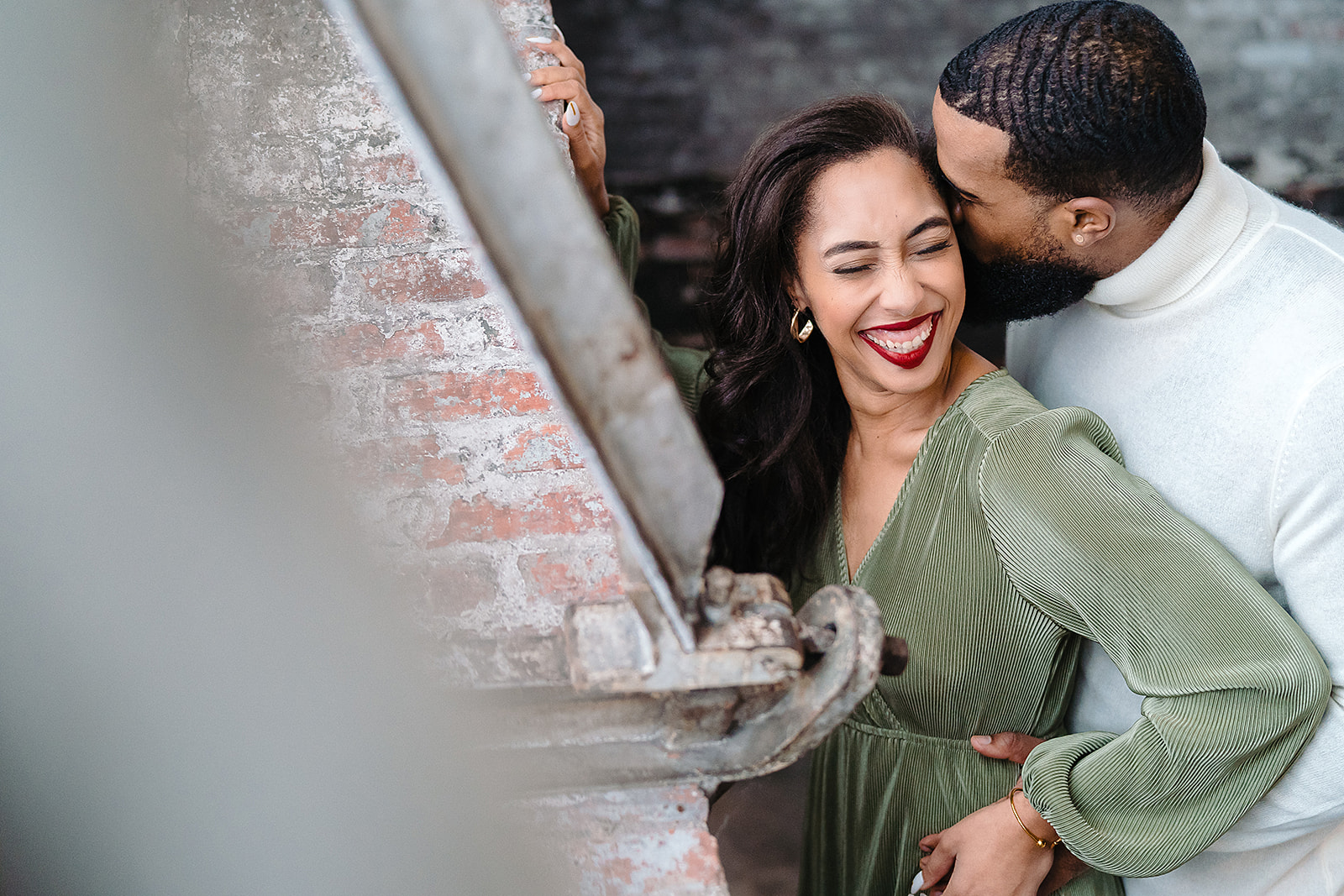 Couple kiss at brick wall in sage dress laughing candid