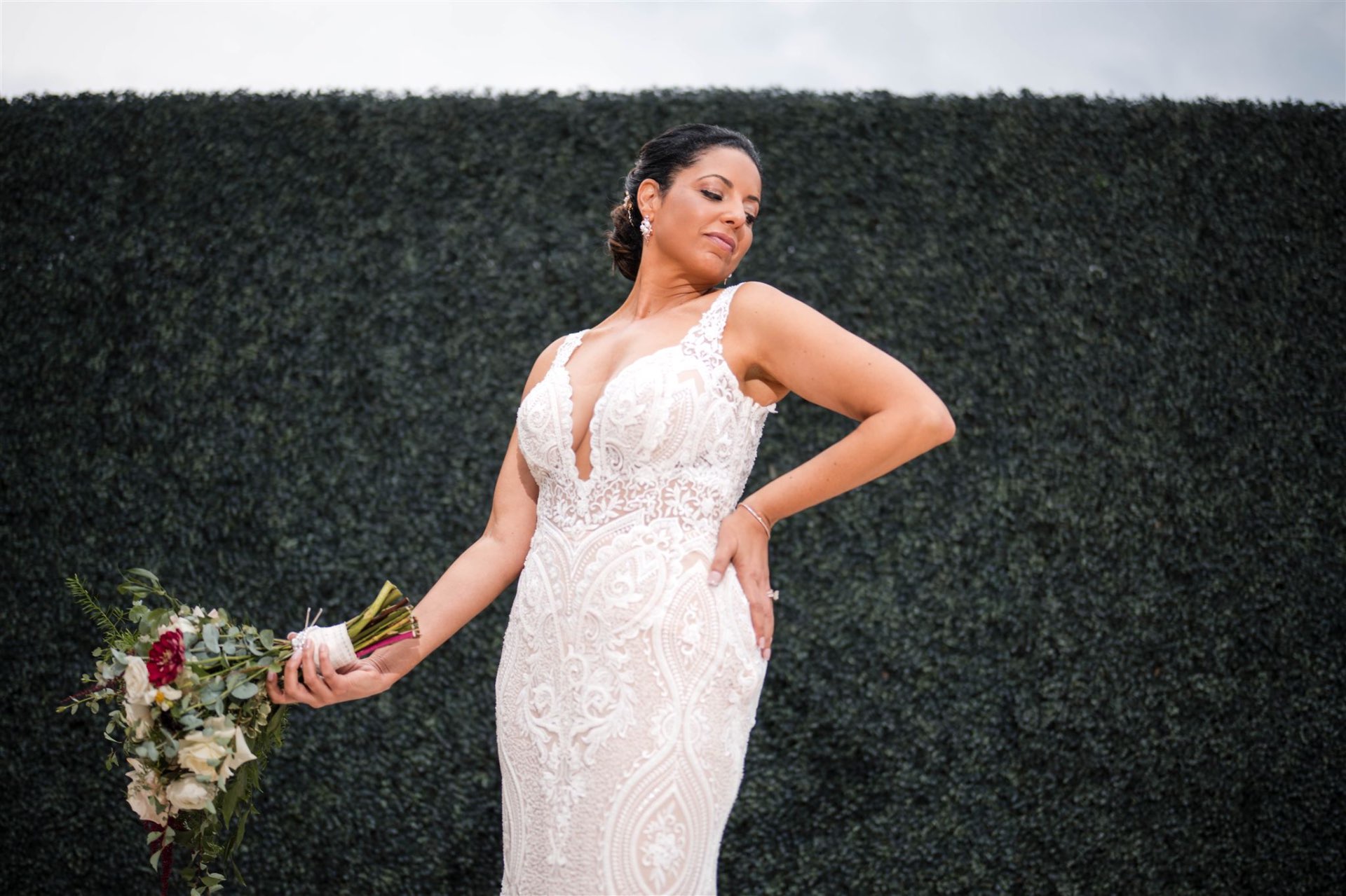 Bride in lace gown posing against hedge wall with bouquet editorial