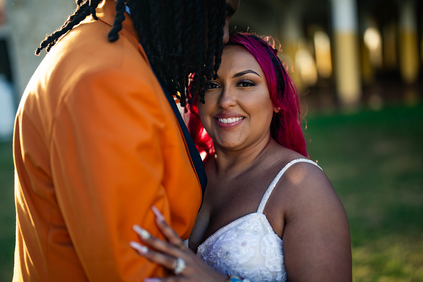 Bride with pink hair smiling at groom in orange jacket with locs intimate