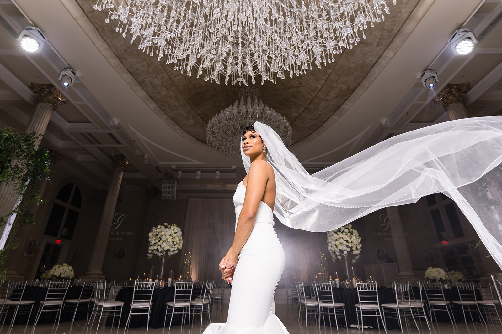 Bride with veil flowing in ballroom under chandelier dramatic