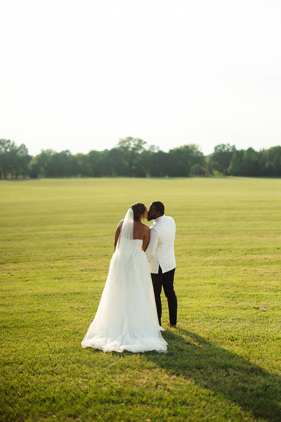 Couple kissing in open field at golden hour with veil and white jacket