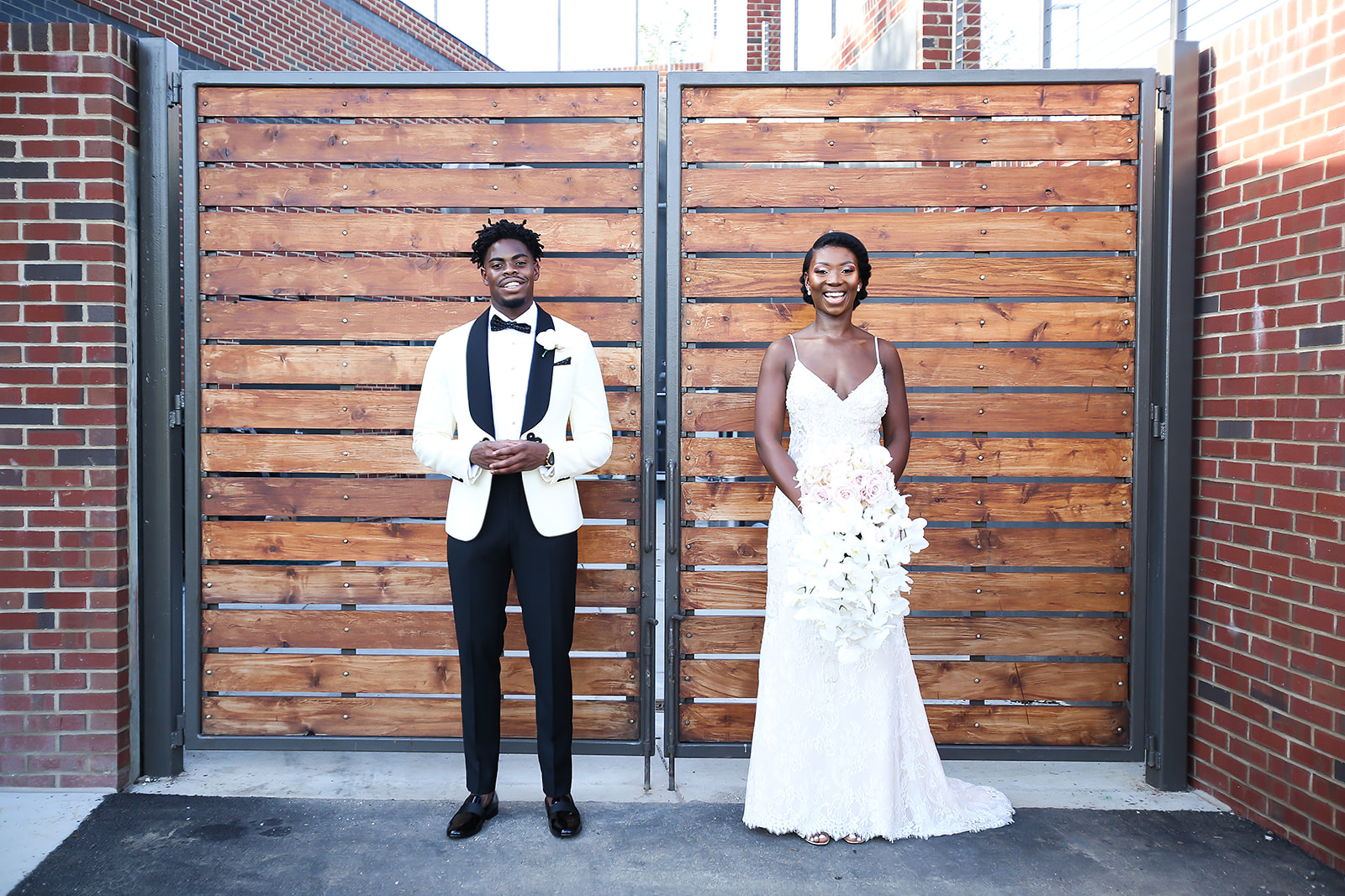Couple standing apart at wood gate brick wall white jacket bouquet editorial