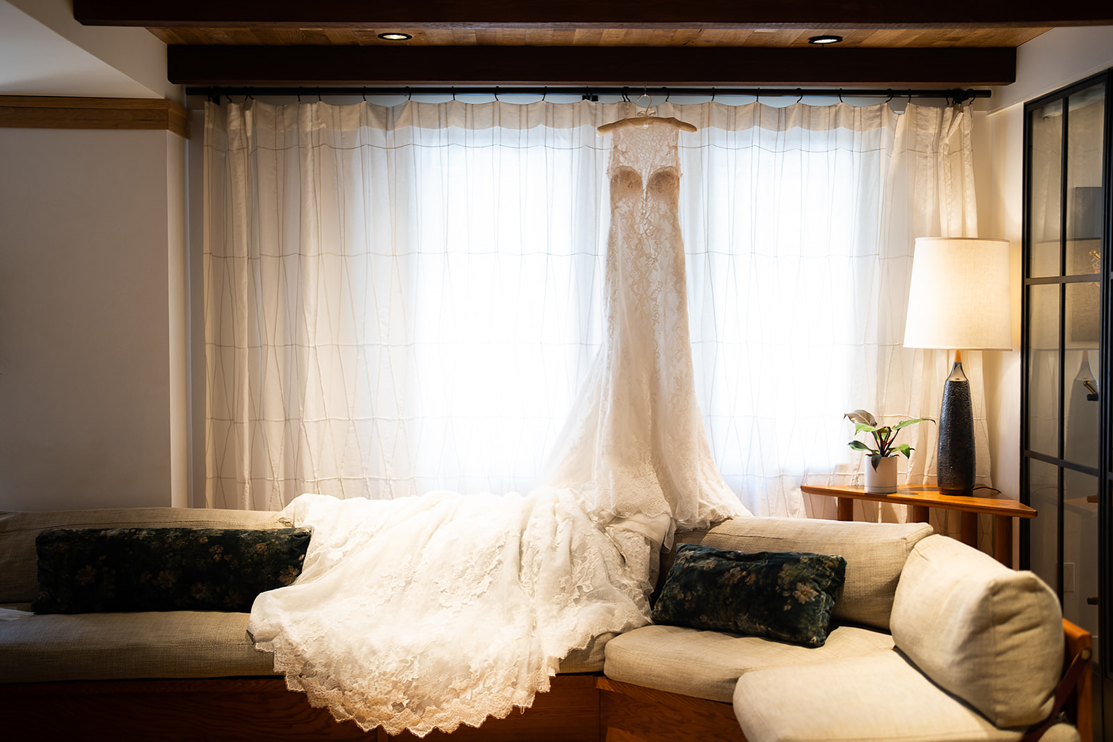 Wedding dress hanging in window with curtains lace on couch warm light