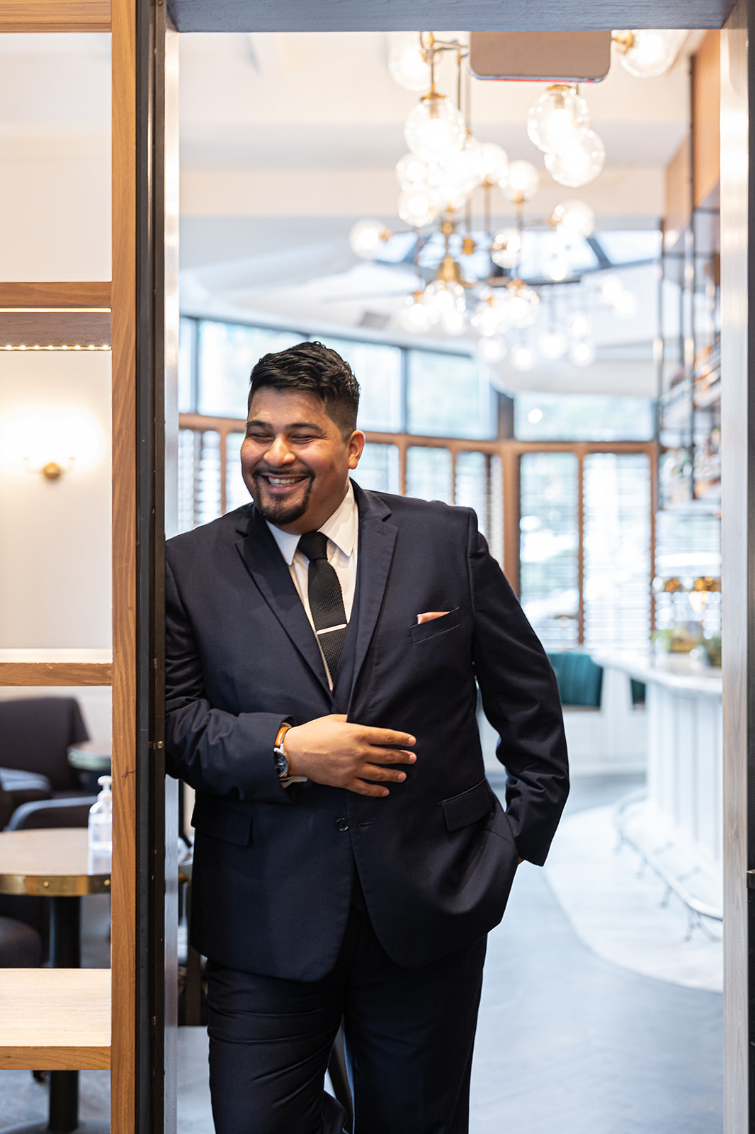 Groom in navy suit laughing in doorway with chandelier at restaurant